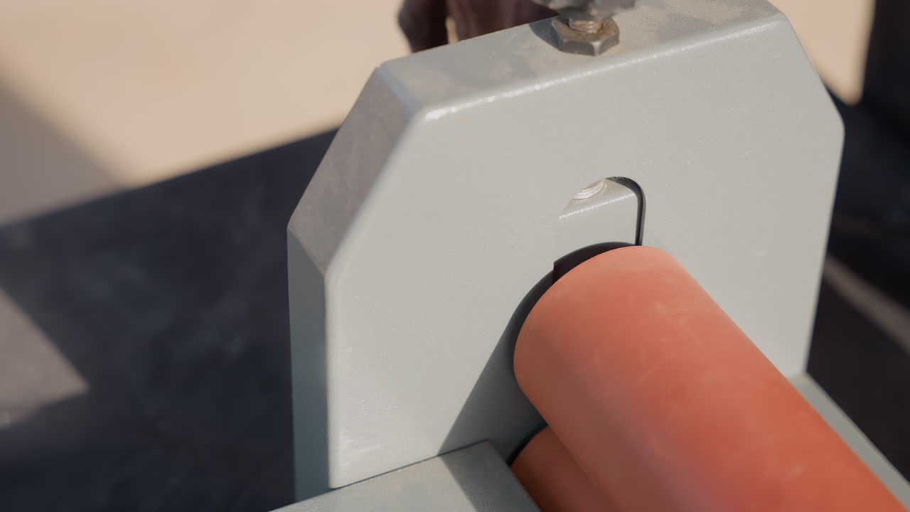 Closeup clamp holding orange rod, black carpenter hand adjusts metal guide, workshop light, dust specks, precise alignment, carpentry setup, tool maintenance, tactile motion, industrial texture