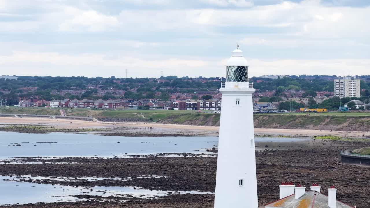 A steady daytime pan reveals St Mary’s Lighthouse against a rocky shoreline and distant town, under overcast skies with soft, diffused lighting
