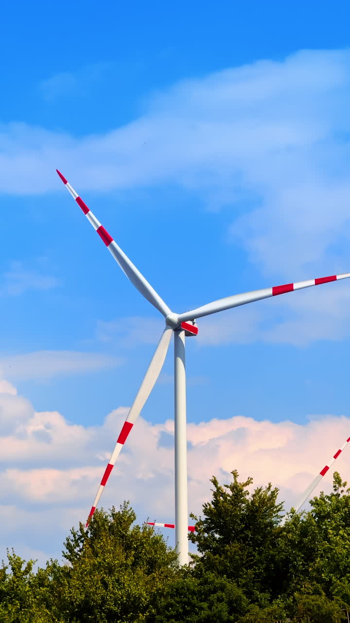 Wind turbine against a clear blue sky. A large wind turbine stands tall among green trees under a bright blue sky with clouds in the background
