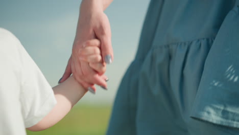 잔디을 가로질러 걸어가는 어린 소년의 부드러운 순간의 클로즈업, 부분적으로 보이는, 웃는, 그는 그의 어머니의 손을 잡고, 파란 드레스를 입고, 그녀의 얼굴을 보이지 않습니다.