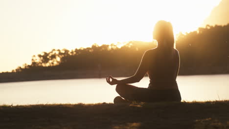 woman meditating doing yoga by beautiful lake and mountains at sunset