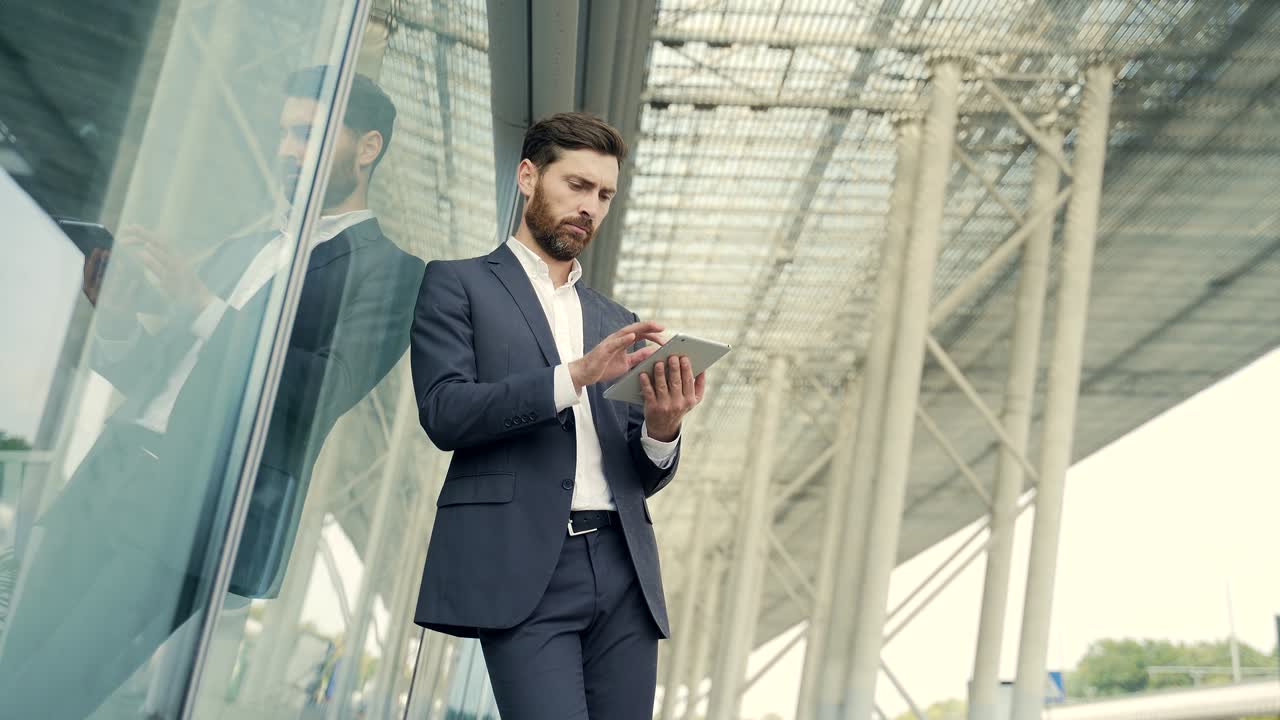 elegante hombre de negocios barbudo en traje de negocios formal de pie trabajando con la tableta en las manos en el fondo edificio de oficinas modernas afuera.
