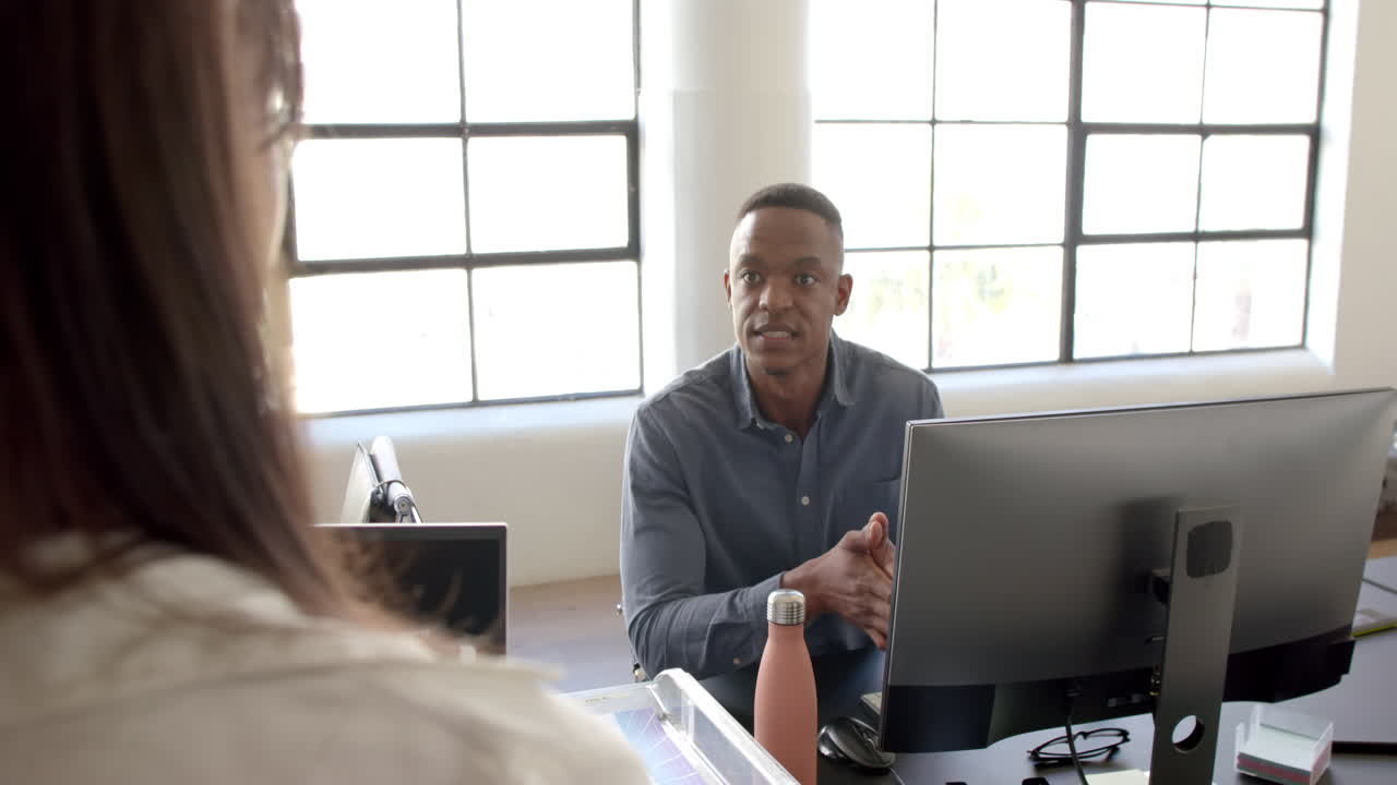 Discussing project details, African American man talking to colleague at desk with computer in offic