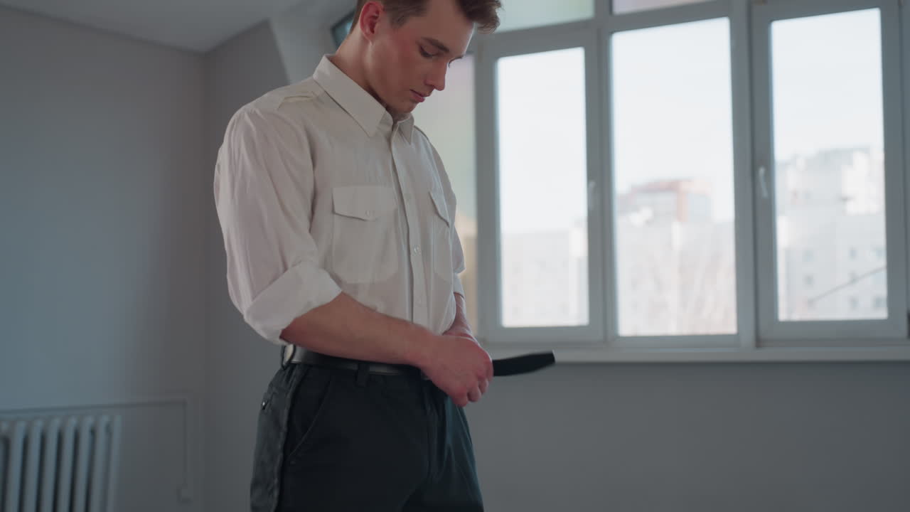 Side profile view of young man fastening belt around waist beside window overlooking urban residential building with antenna outside conveying morning dressing routine focused attention and detail