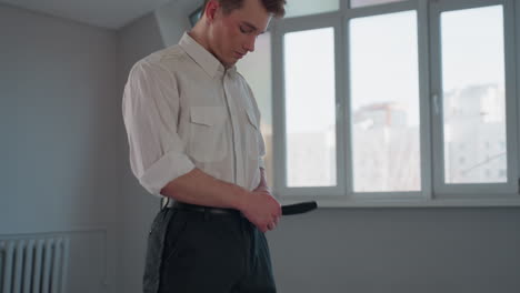 side profile view of young man fastening belt around waist beside window overlooking urban residential building with antenna outside conveying morning dressing routine focused attention and detail