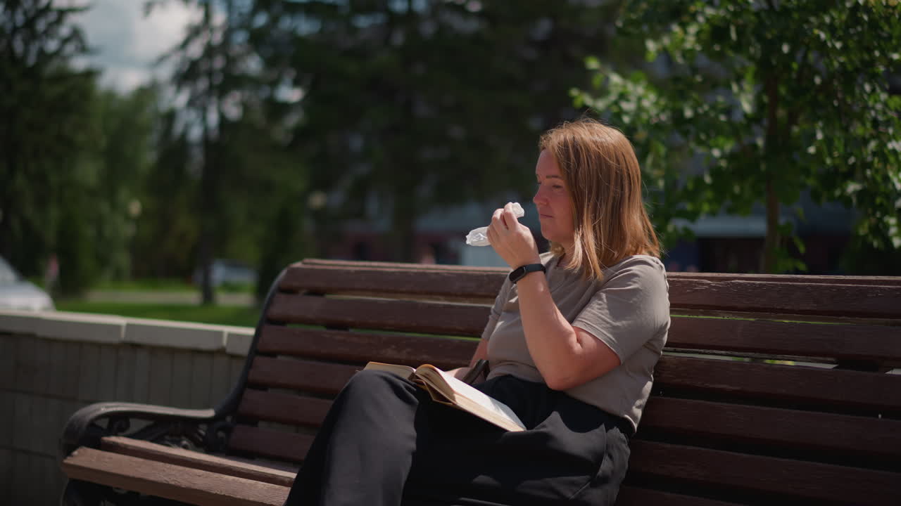 Young lady seated on wooden bench outdoors with open book resting on lap, reaching into pocket to remove napkin while car passes in background and trees sway gently in summer