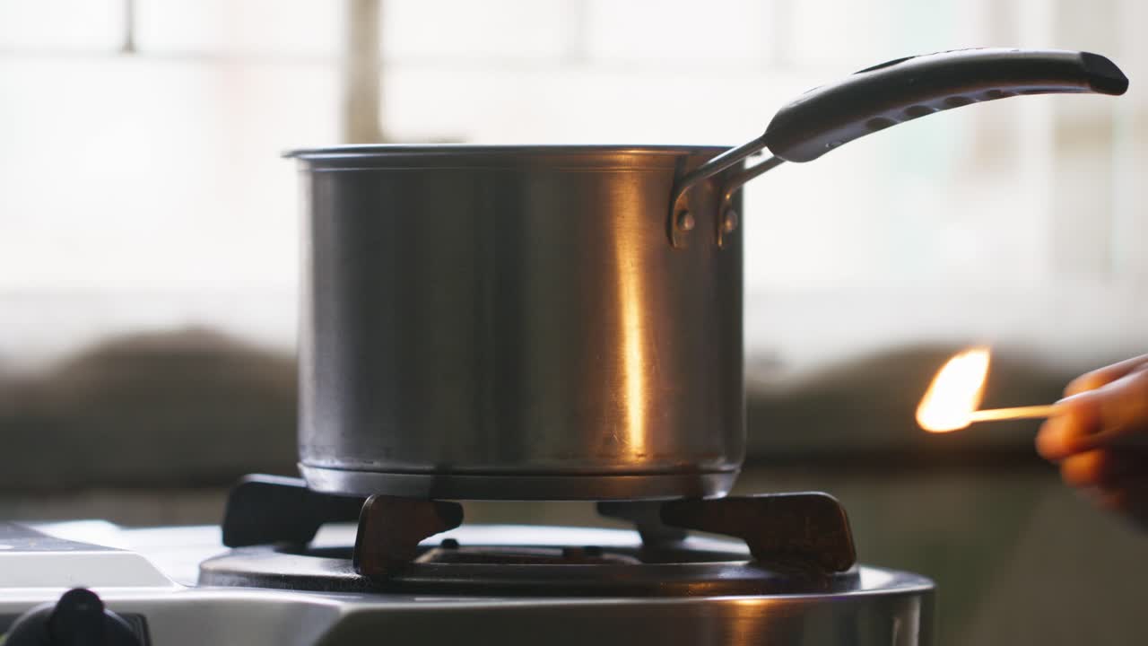 A metal cooking pot on a stovetop and a hand lit the stove with a matchstick