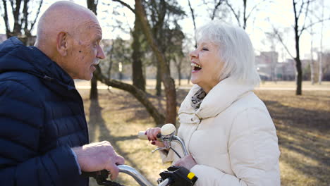 Close-up-view-of-a-senior-couple-talking-and-laughing-in-the-park-on-a-winter-day.-The-man-is-holding-a-bike