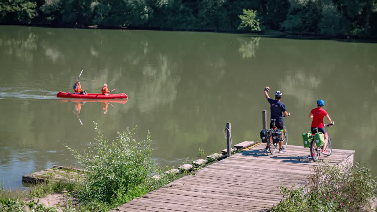 Bikers couple at pier waving to happy friends canoeing on Drava river, Muta