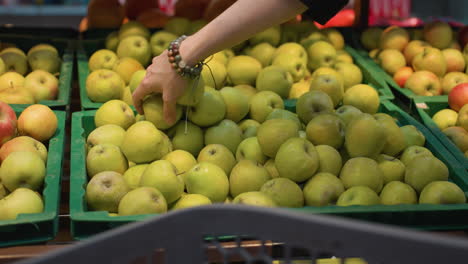 view of a hand wearing bead bracelets picking up apples from a display in a grocery store. the hand is seen selecting and taking two apples. shot with a handheld camera