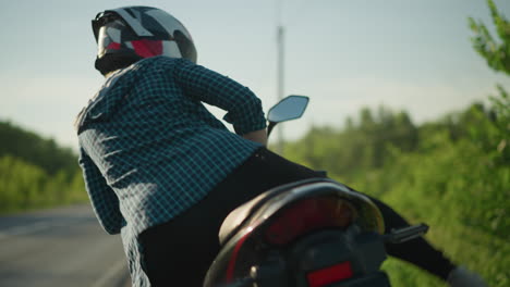 a close-up of a biker dismounting from a motorcycle as she step away, with a blurred view of a car with its headlights on in the background, coming from the distance on the road