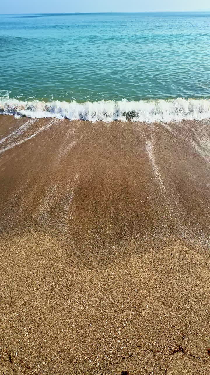 Bølger ruller blidt ind over en sandstrand under en klar blå himmel ved middagstid.