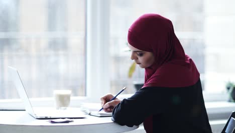 Una-Atractiva-Chica-Musulmana-Está-Escribiendo-Algo-En-Su-Cuaderno-Y-Luego-Comienza-A-Escribir-Algo-En-El-Teclado-De-La-Computadora-Portátil.-Imágenes-En-Cámara-Lenta-En-Interiores