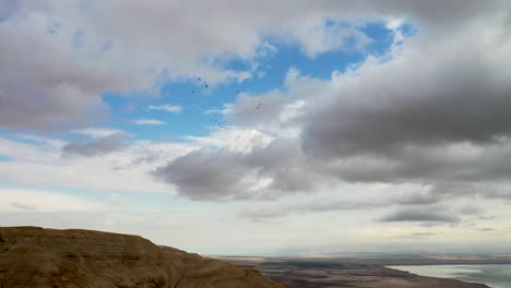 flock of birds fly in rounds over desert mountains, deadsea in the background, cloudy sky, long-shot