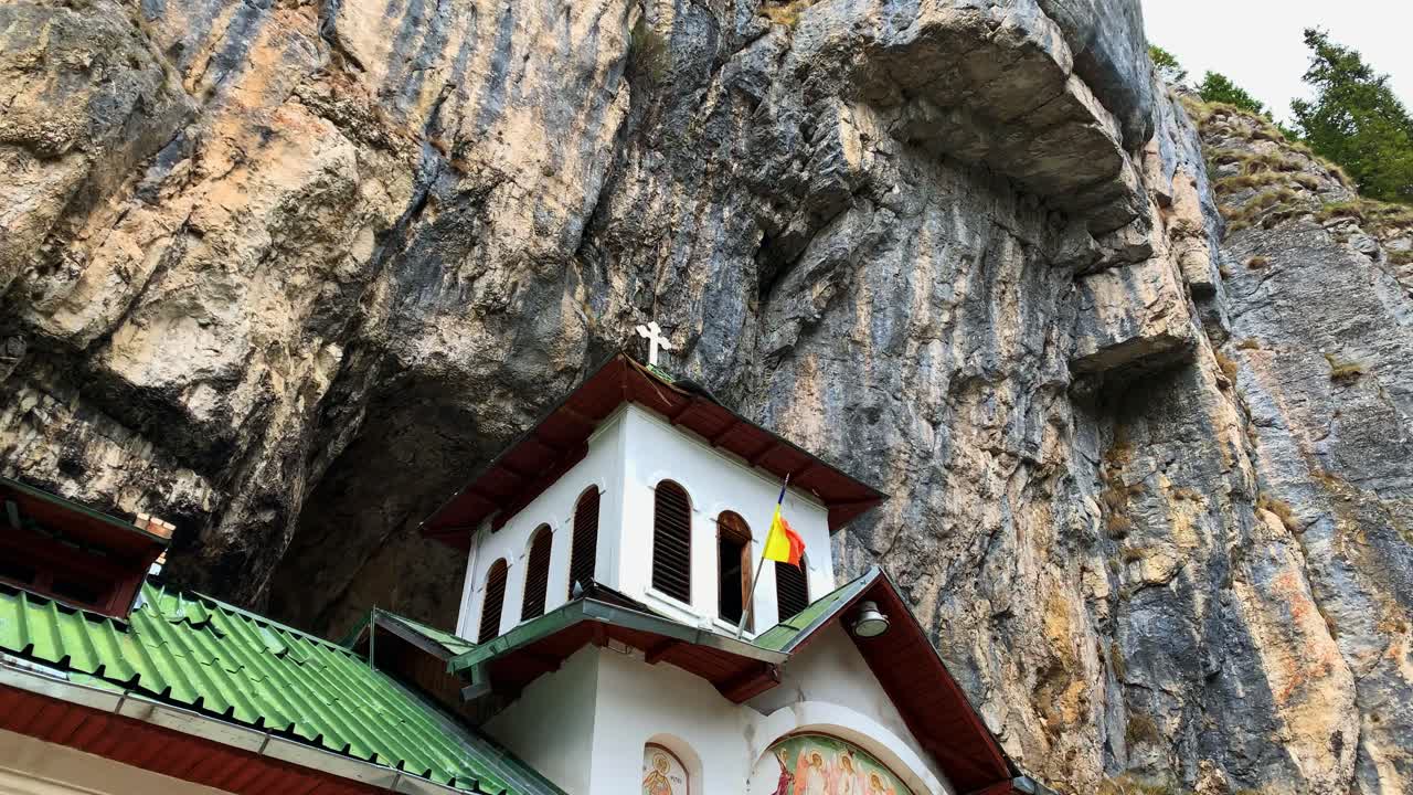 Astonishing View at the Top of Ialomitei Cave Monastery Located in Bucegi Mountains with Romanian Flag on a Windy Day under a Huge Mountain which Protrudes above the Entrance