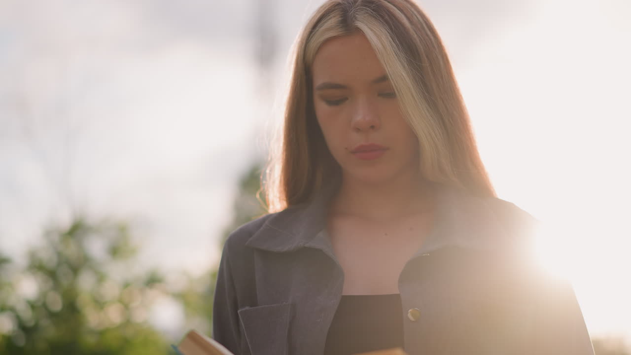 donna in abiti grigi in piedi all'aperto con la testa leggermente inclinata, che gira le pagine di un libro in mano mentre la luce solare si riflette dolcemente intorno a lei a sinistra, sullo sfondo ci sono alberi sfocati
