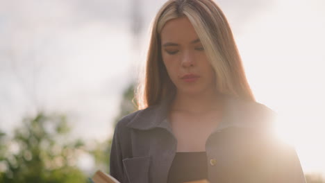 mujer con ropa gris de pie al aire libre con la cabeza ligeramente inclinada, volviendo páginas de un libro en su mano mientras la luz del sol se refleja suavemente a su alrededor a la izquierda, el fondo presenta árboles borrosos