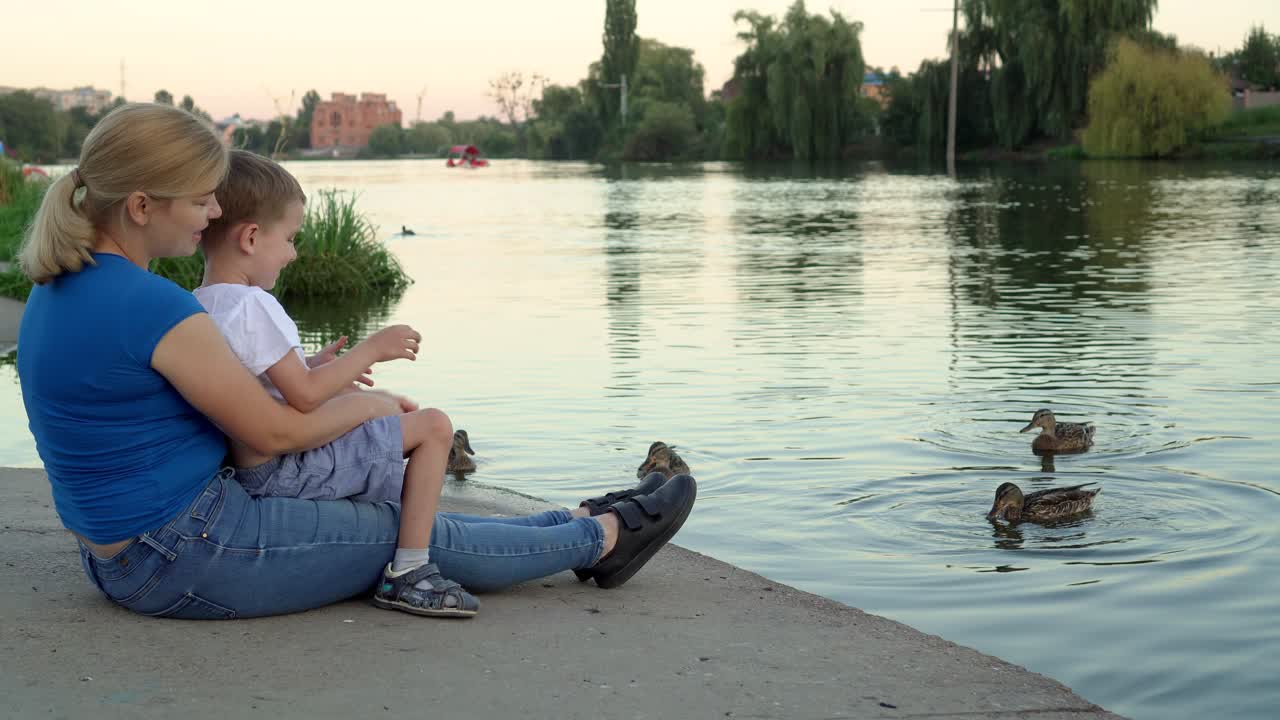 의료 마스크를 입은 어머니와 아이는 호수에서 오리에게 먹이를 주고 있습니다.