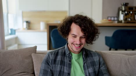 Ein-Glücklicher-Brünetter-Typ-Im-Karierten-Hemd-Mit-Lockigem-Haar-Freut-Sich-über-Seinen-Sieg,-Während-Er-Mit-Einem-Laptop-Auf-Dem-Sofa-In-Einem-Modernen-Apartment-Arbeitet