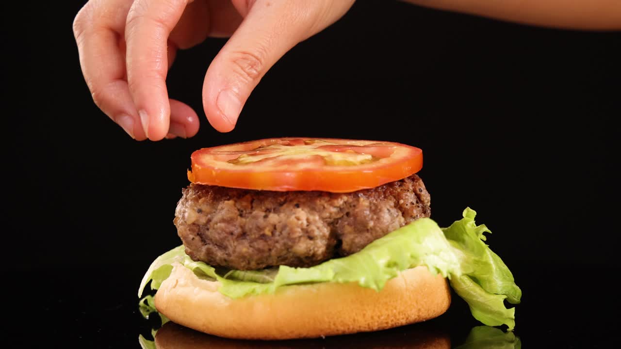 A hand carefully adds a tomato slice onto a beef burger with lettuce and bun, under bright studio lighting against a black background, using close-up framing