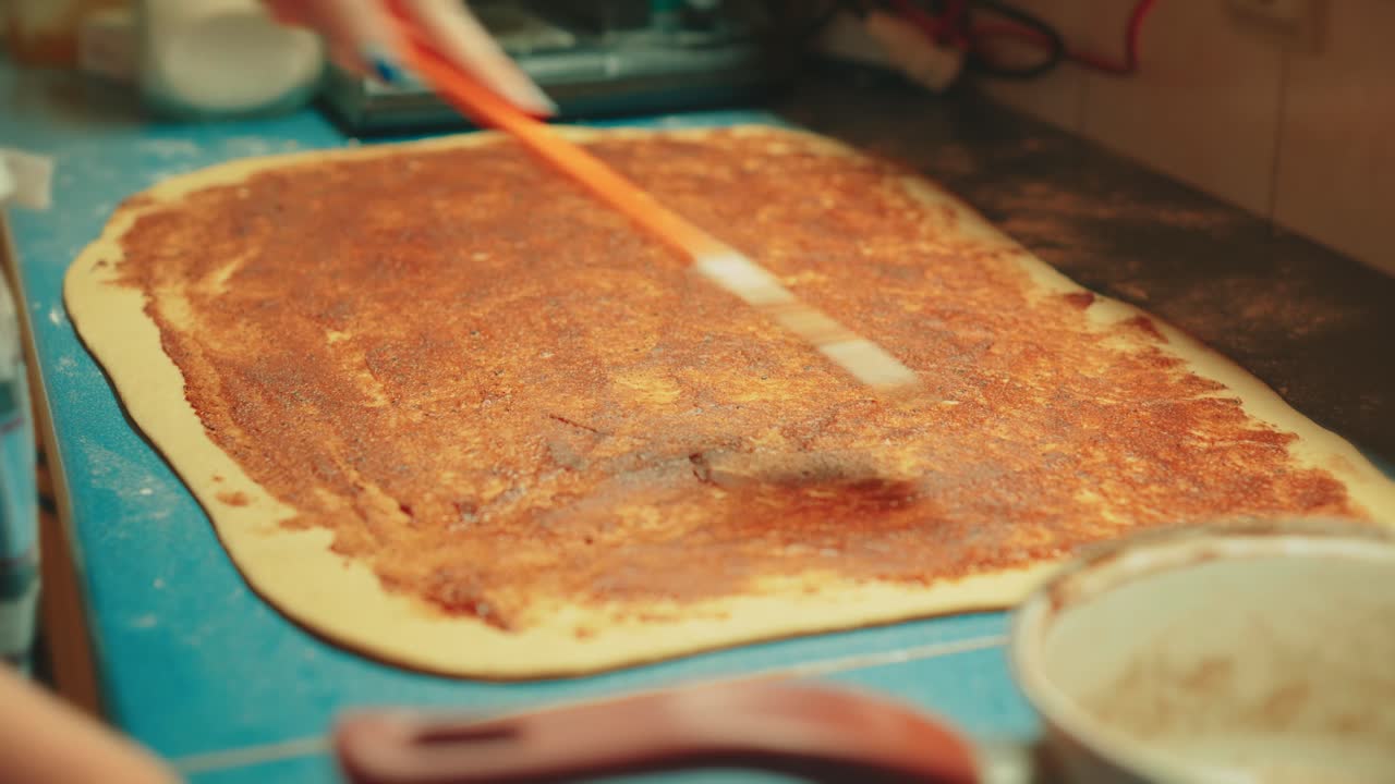 Close up of teenage hands using a spatula to spread a mixture of cinnamon, sugar, and butter on rolled out dough. Rich texture of sweet filling for traditional cinnamon buns