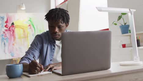 Afroamerikanischer-Männlicher-Künstler-Mit-Laptop,-Der-Sich-Notizen-Macht,-Während-Er-Auf-Seinem-Schreibtisch-Im-Kunststudio-Sitzt