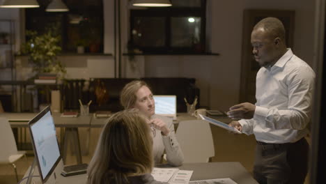 american man employee holding a tablet and explaining a project to female coworkers who are sitting at desk in the office 1