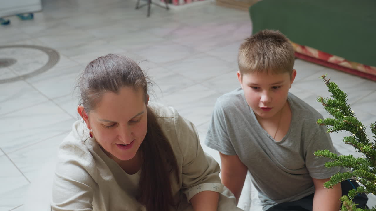 Woman and son sitting together in casual wear, opening box near Christmas tree decorated with glowing lights, festive cozy indoor atmosphere capturing joyful family holiday preparation moment