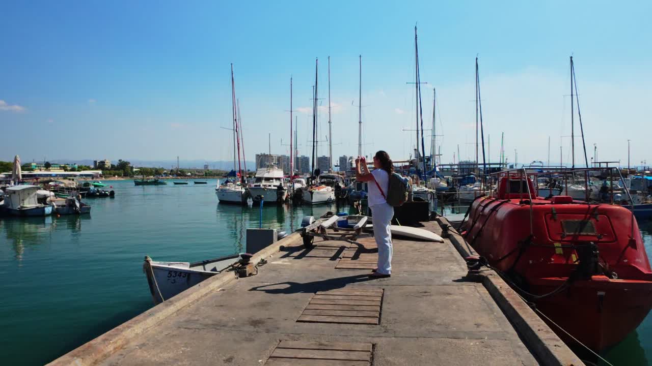 Tourist woman photographing sailboat masts in marina with smartphone