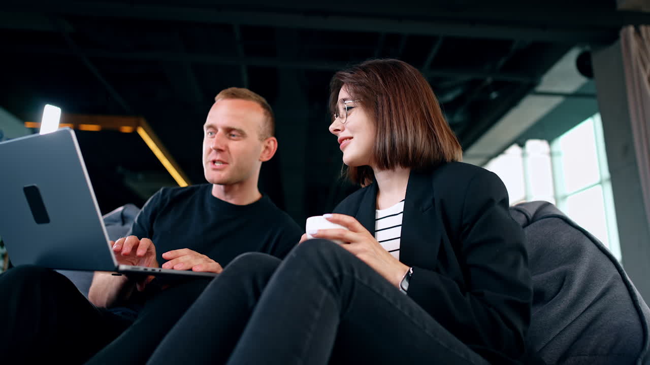 Young people work sitting in the soft chairs. Man and woman use laptop looking on the screen and discussing. Low angle view.