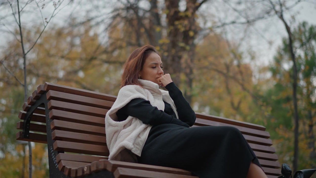 Woman rests on wooden bench in quiet park, wrapped in scarf and dark coat, eyes closed and hand near cheek while leaning back in calm, reflective pose amid muted autumn trees and fallen leaves