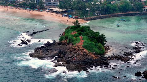 a tropical rocky islet off sri lanka’s mirissa coast rises with lush greenery, surrounded by crashing waves, facing a golden sandy beach lined with palm trees and vibrant coastal life.