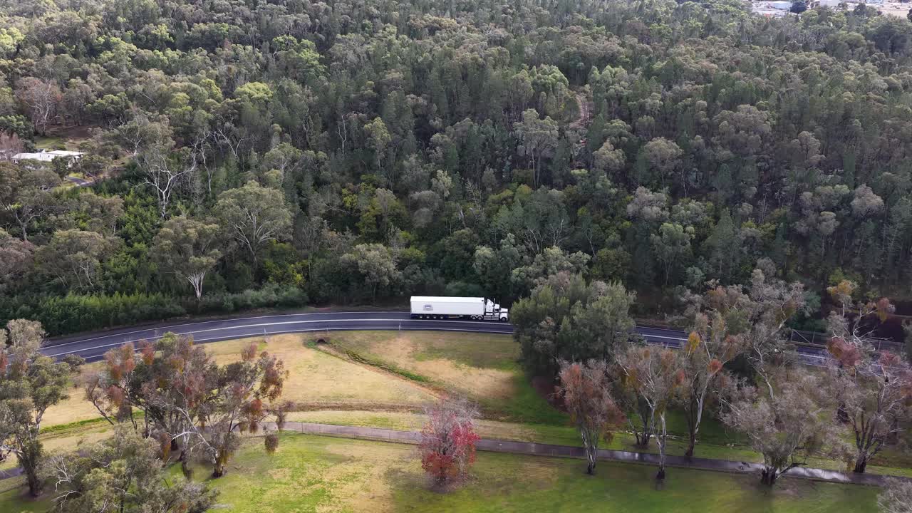 A white semi truck travels along a winding countryside road bordered by forest and open fields, captured in smooth daylight aerial footage near Coonabarabran, NSW