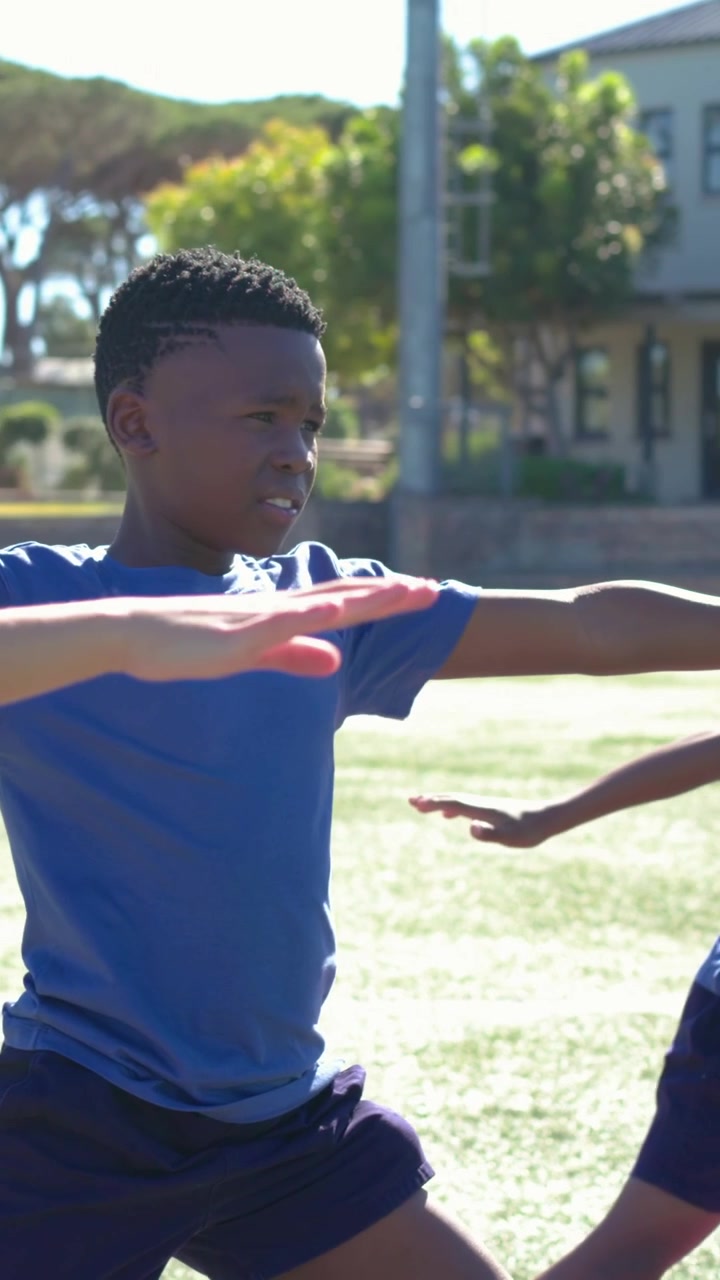 Vertical video: Extending right arm, boy in blue shirt holding wide stance on field starting warmup