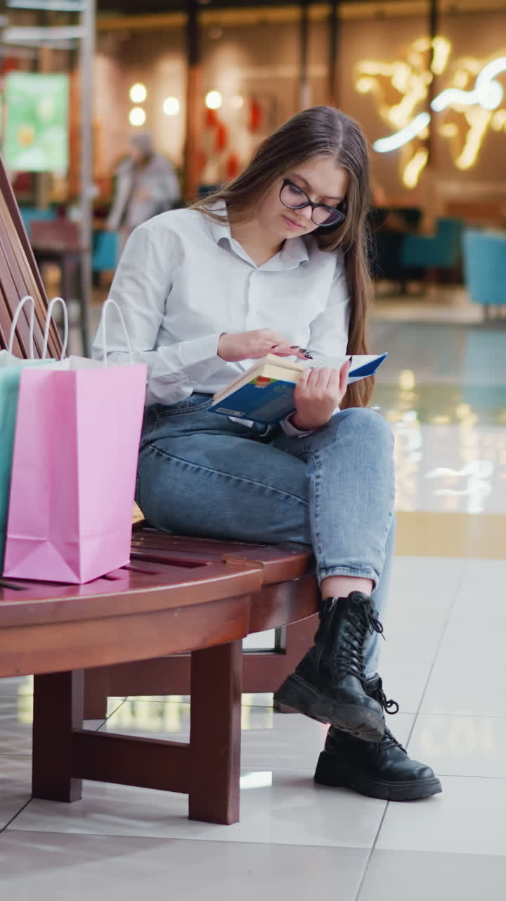 novelista sentado en un banco de madera moderno, piernas cruzadas, leyendo un libro con bolsas de compras cerca, en un centro comercial con carteles y otros compradores en el fondo