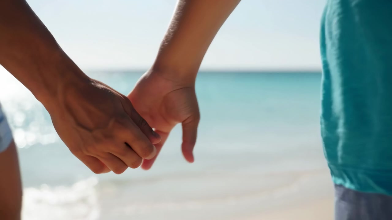Waist-to-hands close-up of an adult walking along a sandy shoreline, their linked hands centered lower right with sunlight reflecting off calm ocean