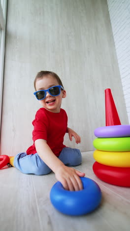 adorable funny toddler boy in red t-shirt and sunglasses sits on the floor. joyful kid plays with toy pyramid. low angle view. vertical video.