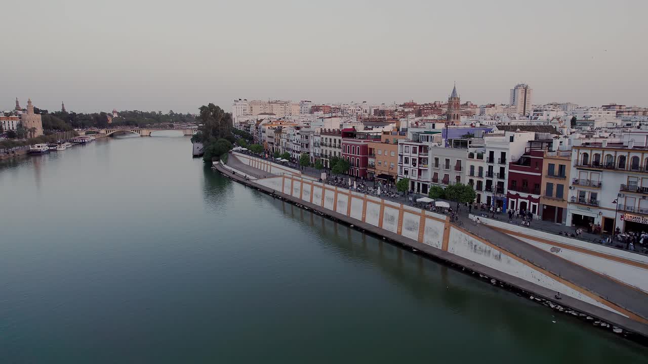 Guadalquivir River separating Triana district on left from Paseo de Cristóbal Colón embankment on right, with Torre del Oro and Puente de Triana in background of Seville cityscape, low aerial, Spain