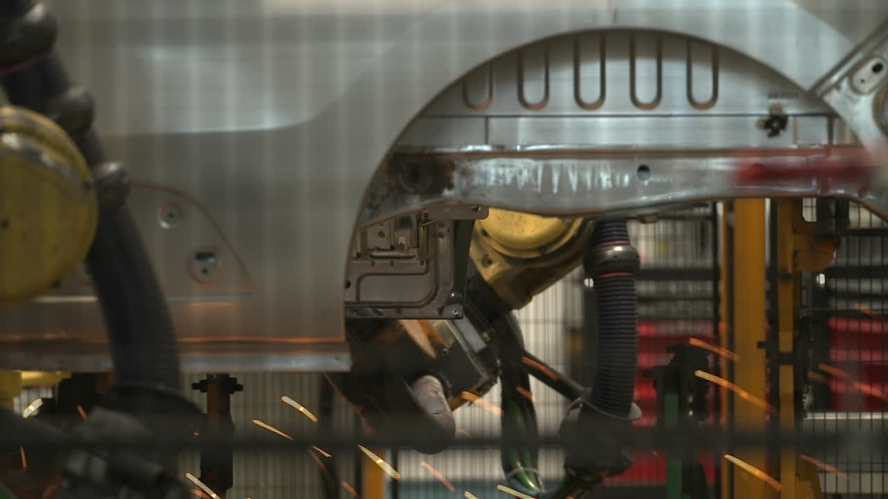 "Inside a bustling car manufacturing plant, robotic arms and skilled workers assemble vehicles on the production line with precision and speed, surrounded by conveyor belts and high-tech machinery."