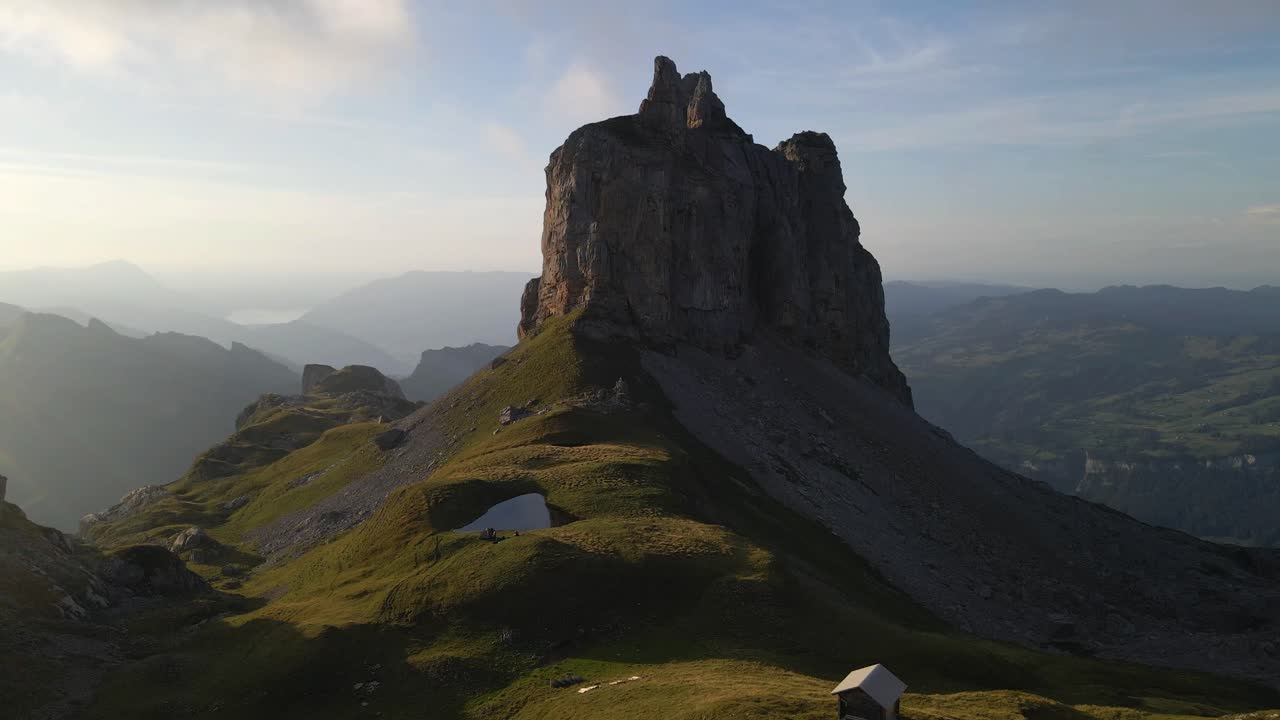 A drone captures a breathtaking aerial view of the Gross Achslenstock, showcasing its rugged, towering rock formations and the surrounding lush green landscape bathed in soft sunlight