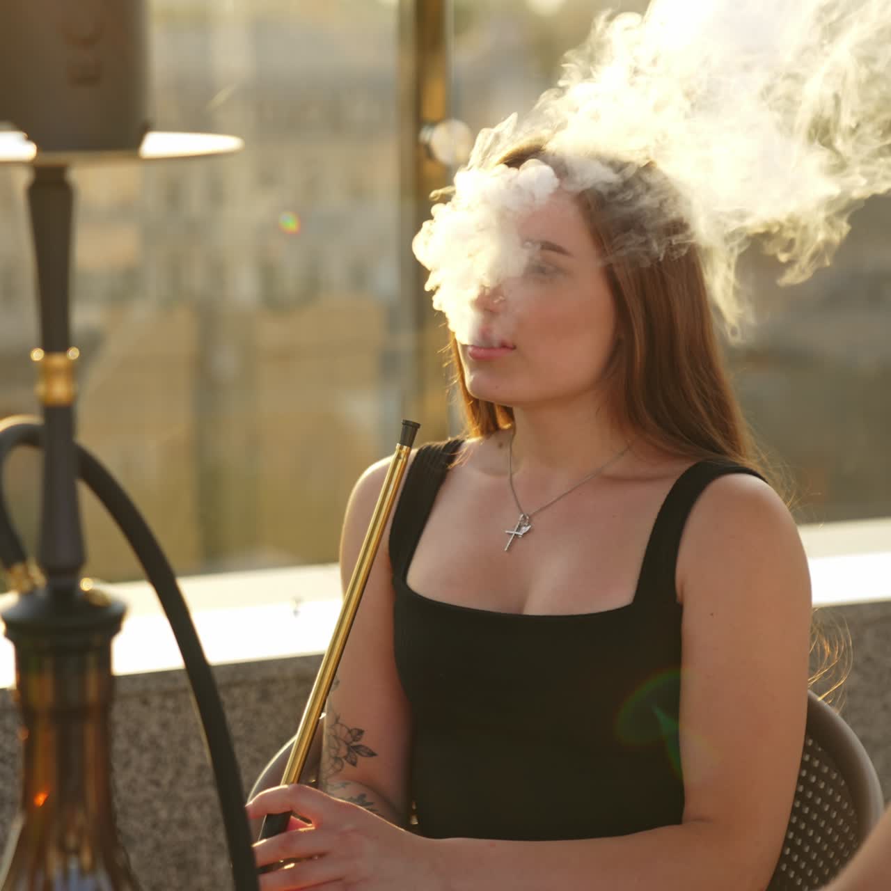 Attractive long-haired lady smoking hookah producing much smoke. Happy girl friends having good time together at summer vacation