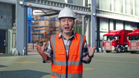 asian male engineer with safety helmet smiling to camera and saying wow while standing , outside of logistics distributions warehouse