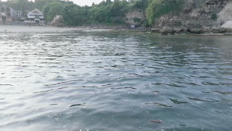 Low-angle-panning-down-drone-shot-of-polluted-water-filled-with-plastic-trash-and-dead-coral-reef-in-the-turqouise-tropical-water-of-Bali-Indonesia