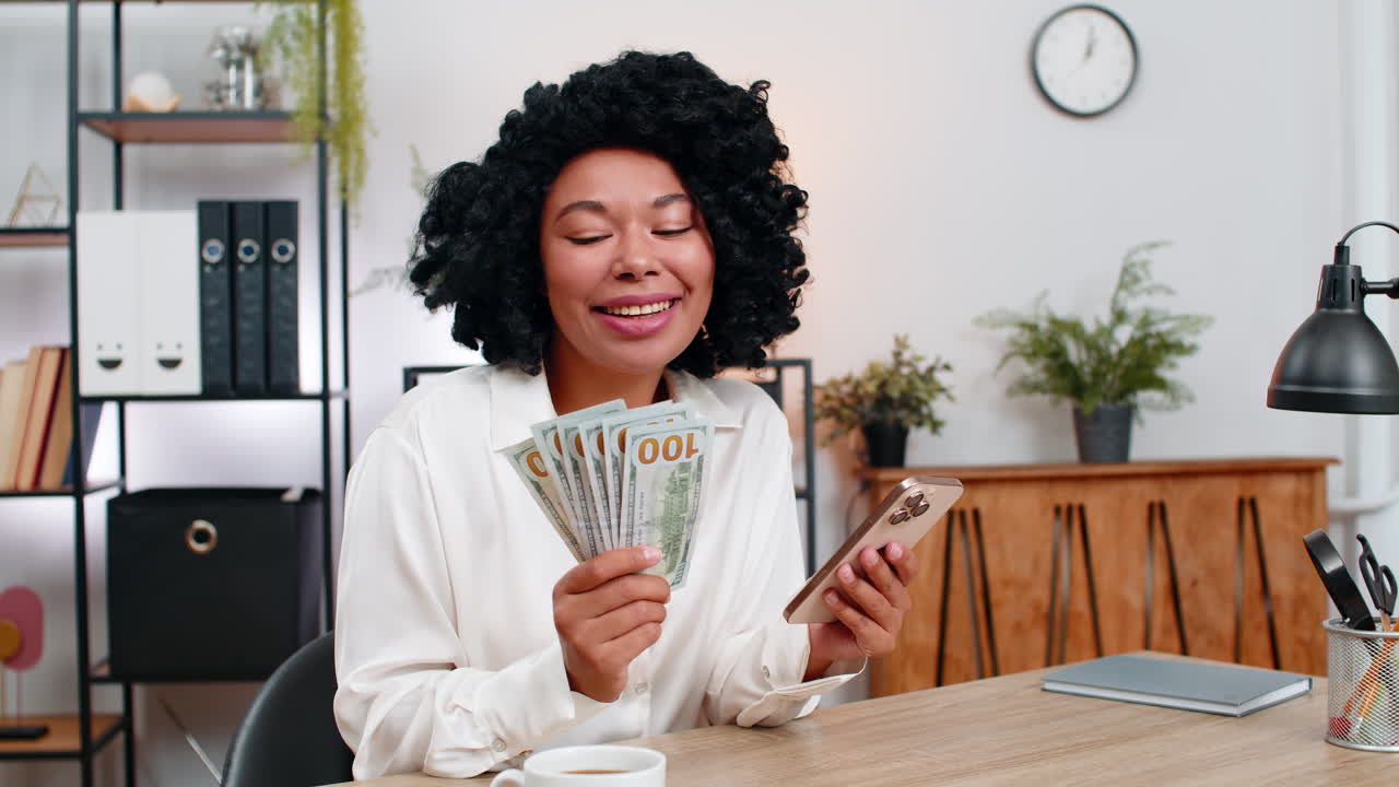 African american businesswoman surprised counts cash holding smartphone planning spending big profit