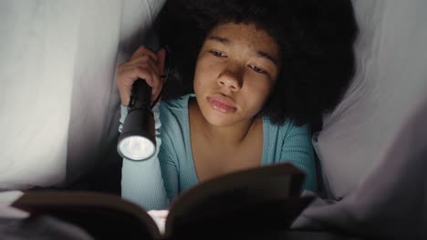 teenage girl spending night time under blanket on reading book with torch