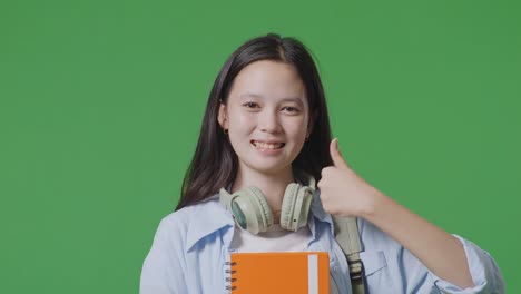 close up de estudiante adolescente asiática con una mochila y algunos libros sonriendo y mostrando pulgares hacia arriba gesto a la cámara mientras está de pie en la pantalla verde estudio de fondo