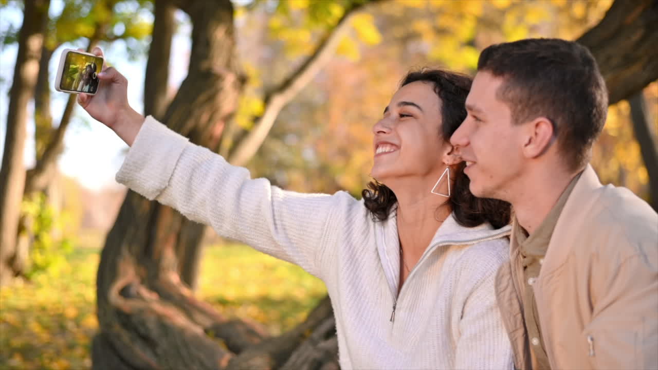 Um casal romântico em um parque no outono. Sorrindo e tirando selfies. Clima de outono, árvores e folhas amareladas ao redor. Câmera lenta