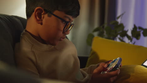 Young-Boy-Sitting-On-Sofa-At-Home-Playing-Game-Or-Streaming-Onto-Handheld-Gaming-Device-At-Night-4