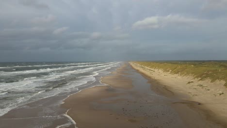 a drone flies at high altitude along the hvide sande beach, capturing wide views of the stormy danish coastline, gray skies, and turbulent sea under a moody evening sky with the dunes to the right