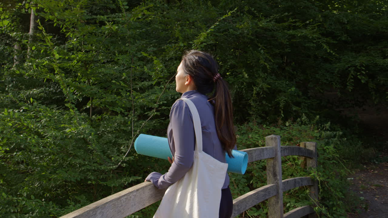 운동복을 입고 요가 매트를 들고 있는 여성이 숲 속의 다리 위에 서서 자연의 평화와 아름다움을 즐기고 있다.